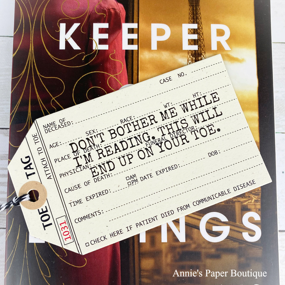 A novelty bookmark tag styled like a vintage medical death tag, printed with humorous and creepy text for readers. The tag is shown on a styled flat-lay background featuring a book and a white wood surface.