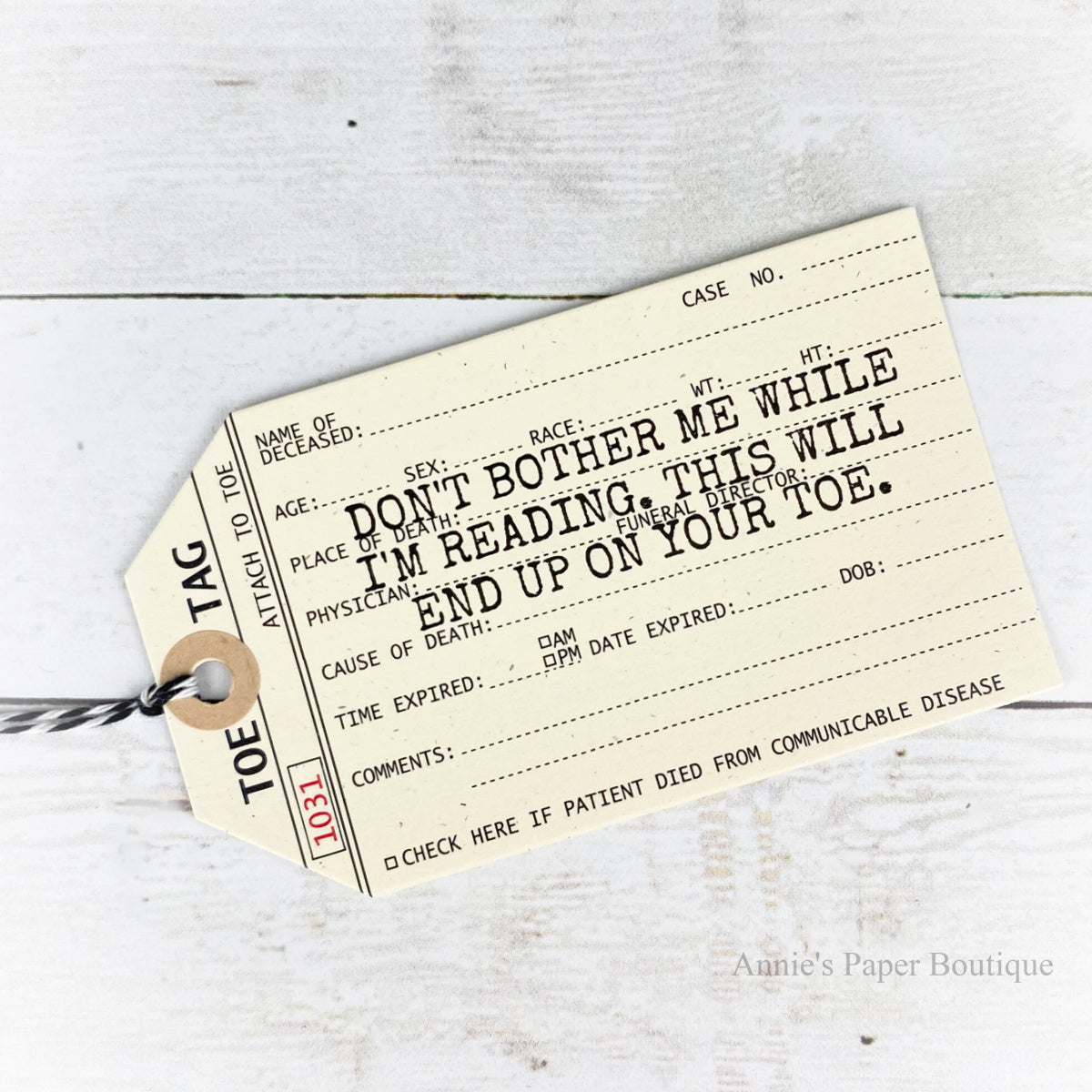 A novelty bookmark styled like a vintage medical death tag, printed with humorous and creepy text for readers. The tag is shown on a styled flat-lay background featuring a book and a white wood surface.