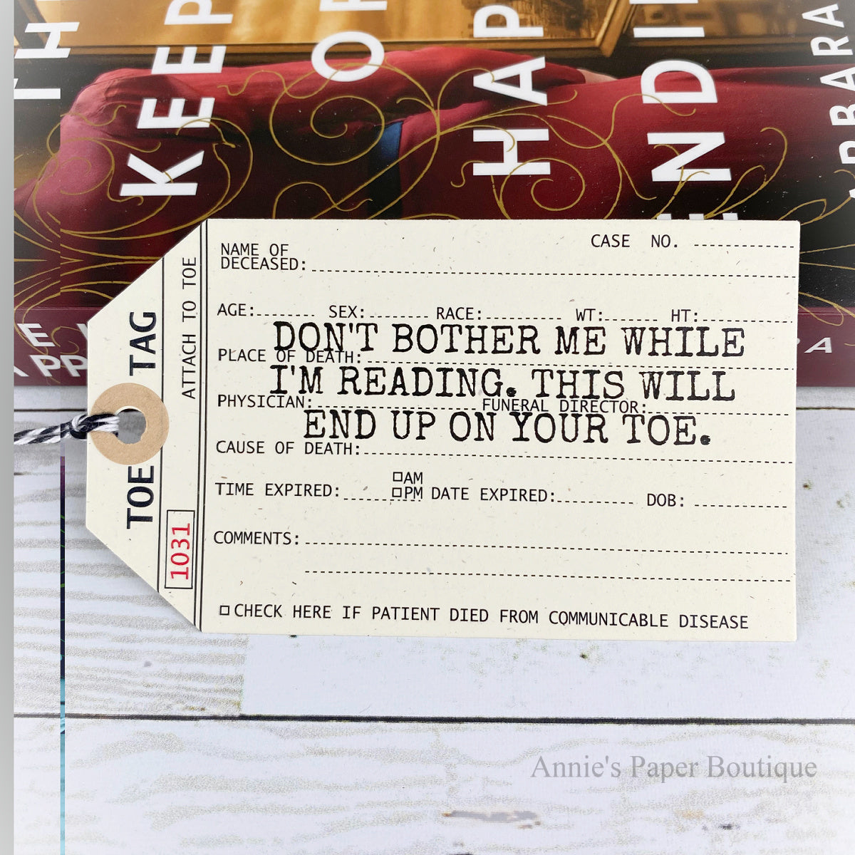 A novelty bookmark styled like a vintage medical death tag, printed with humorous and creepy text for readers. The tag is shown on a styled flat-lay background featuring a book and a white wood surface.